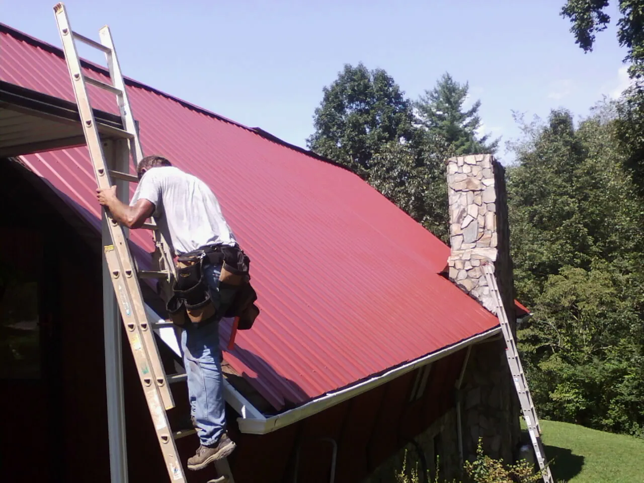 Roofer on a ladder beside a red metal roof and stone chimney — Soffit & Fascia Repair by Roofing Brothers in Glendale